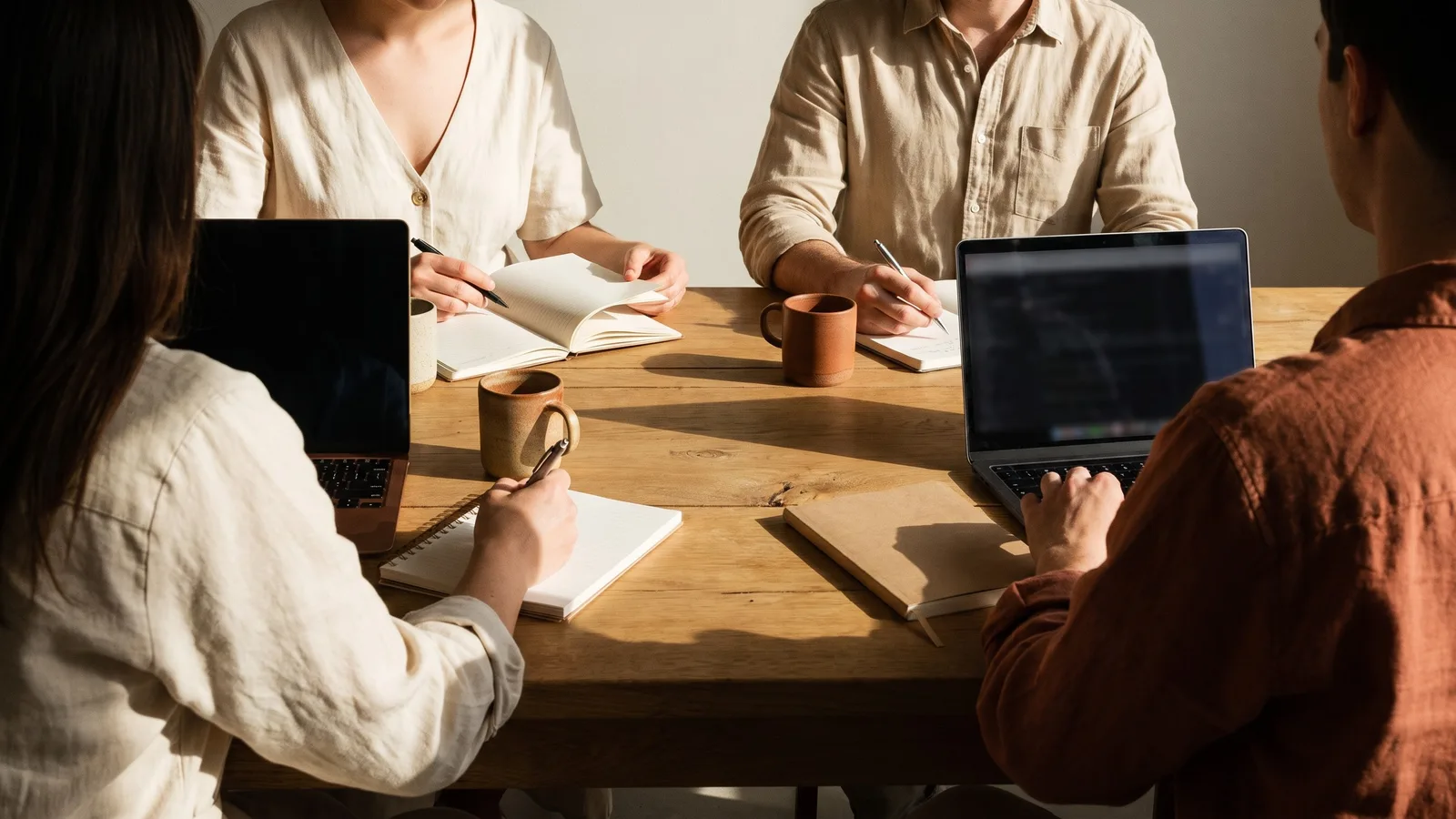 Vier Personen sitzen an einem Holztisch, nur Oberkörper und Hände sichtbar, mit Notizbüchern, Laptops und Tassen im warmen Licht. (KI-generiert)